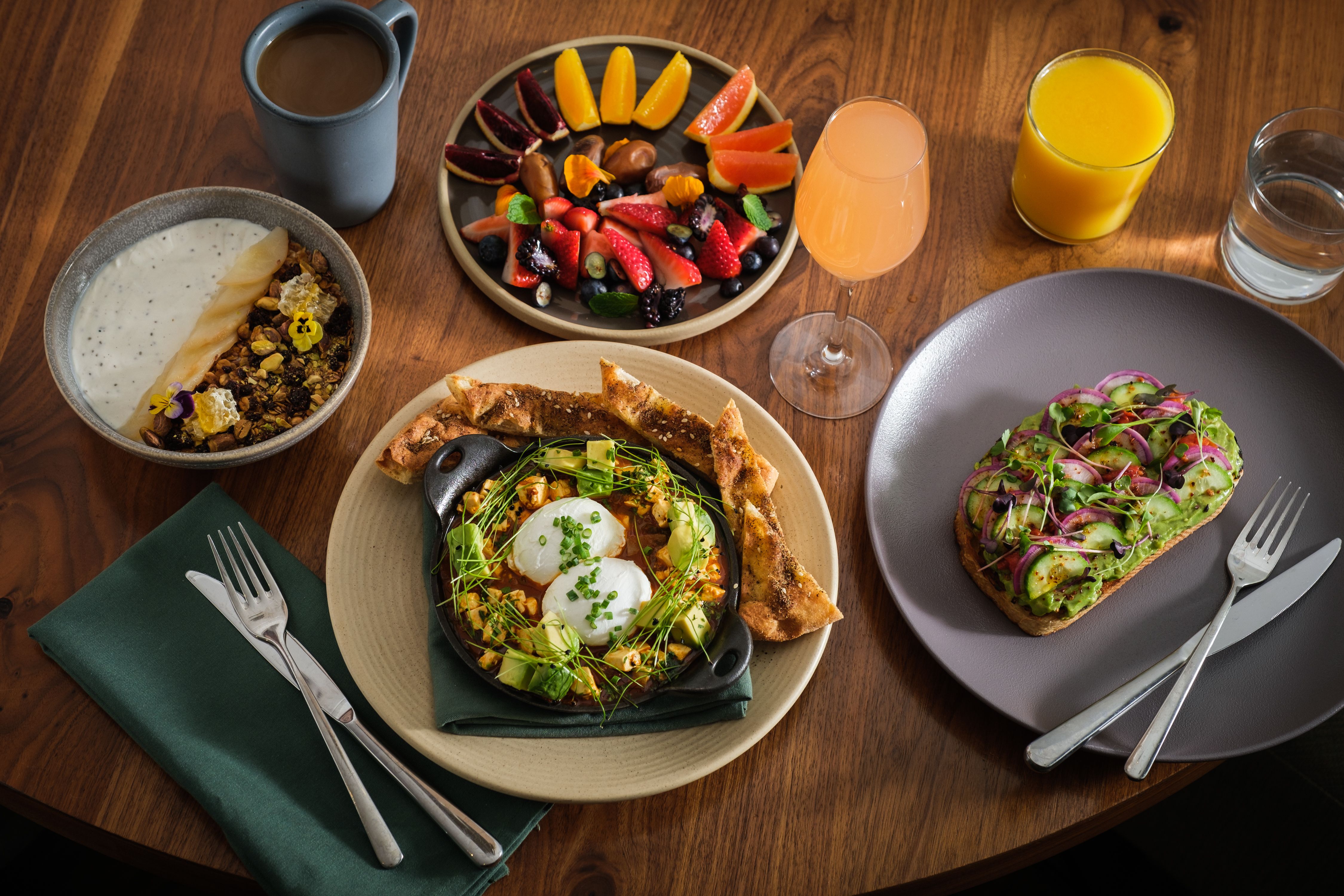 Colorful array of healthy breakfast items including avocado toast, and a platter of fresh fruit displayed on a wood table